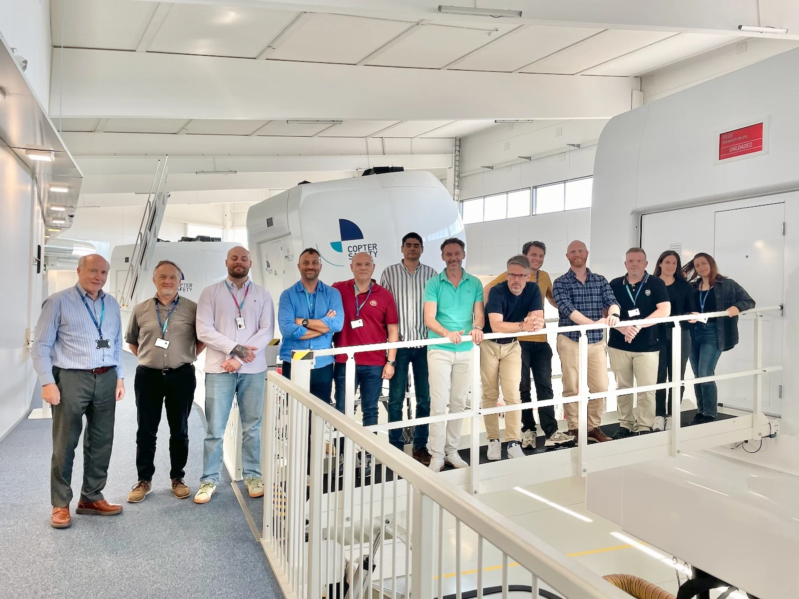 The July 2026 TTI FSTD Evaluators Course cohort on the gantry beside an H125 full-flight simulator at CopterSafety, Helsinki